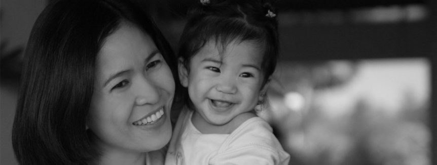 A woman of Asian descent holds her daughter in her arms, both smiling.